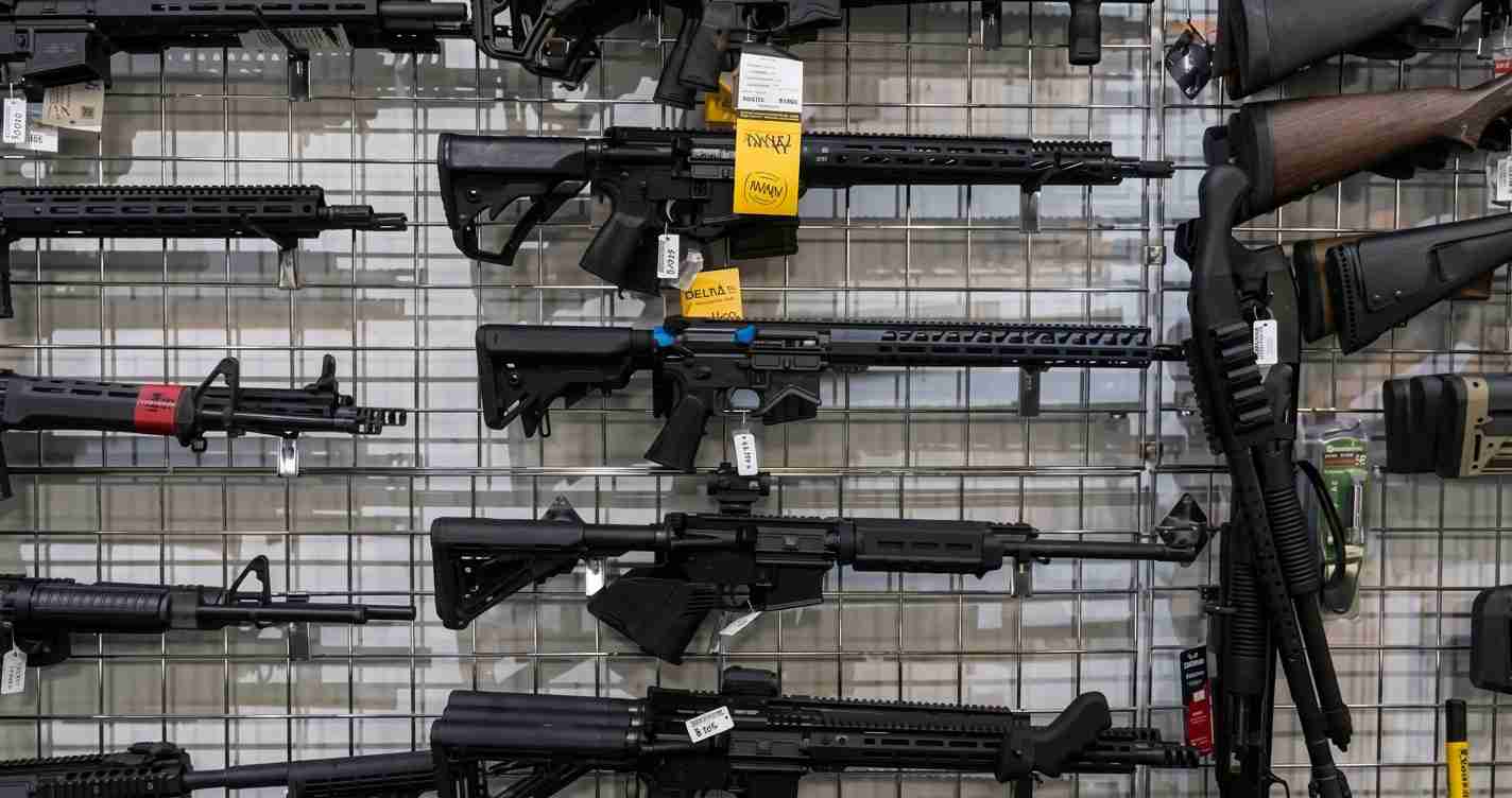 Rack of black rifles displayed on a grid wall in a gun shop, with price tags hanging from some barrels.