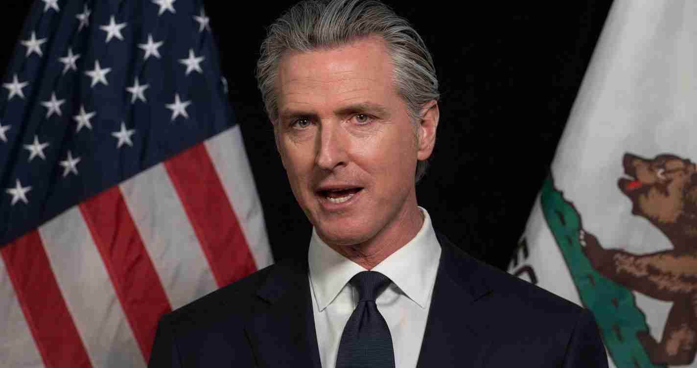 Man in a dark suit and tie speaks, with American and state flags behind him.