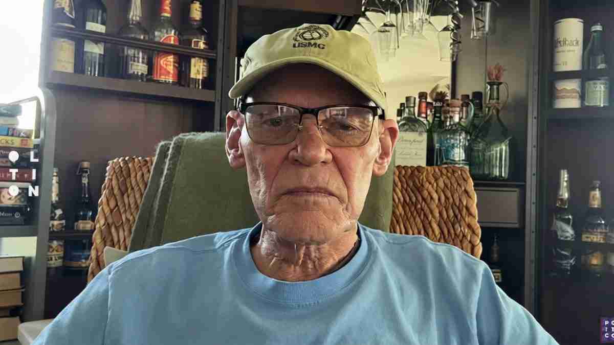 Older man wearing a USMC cap and glasses sits in a home bar with bottles behind him.