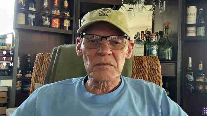 Older man wearing a USMC cap and glasses sits in a home bar with bottles behind him.