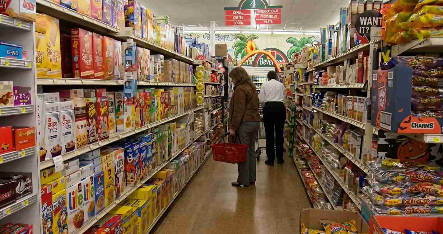 Shoppers browse a cereal aisle in a grocery store; a woman in a brown jacket holds a red basket while a man stands farther back by shelves.
