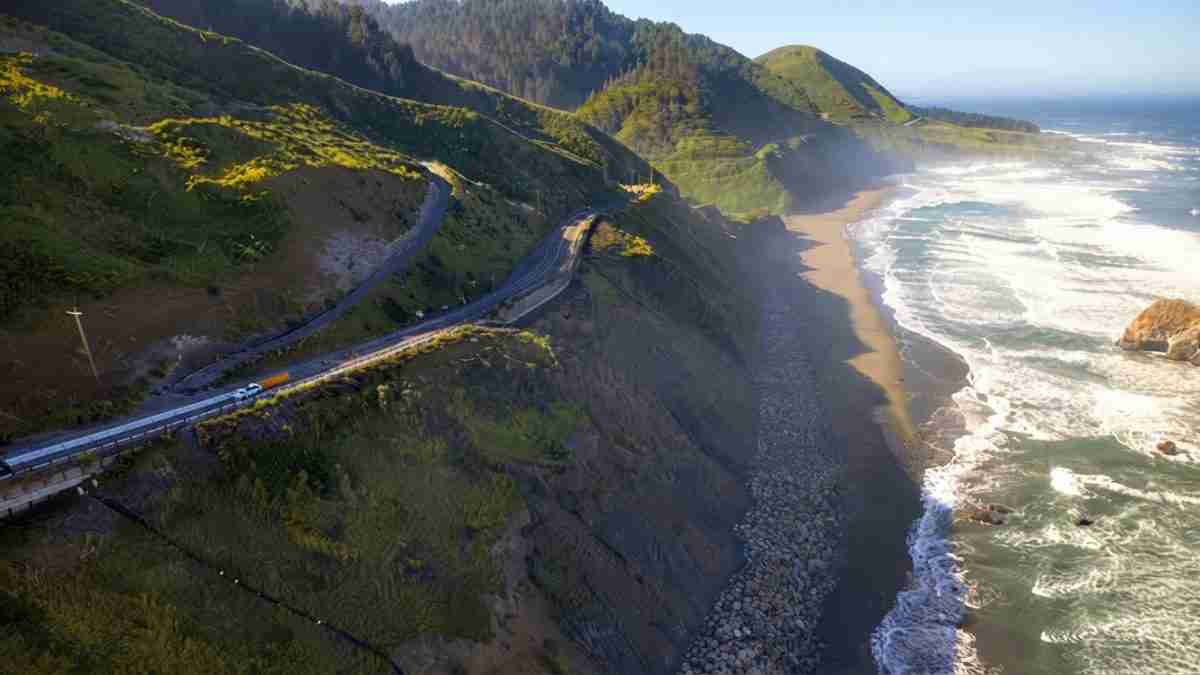 Winding coastal road along steep, green cliffs above a rocky beach and the ocean.