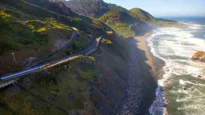 Winding coastal road along steep, green cliffs above a rocky beach and the ocean.