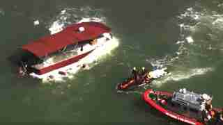 Red overturned passenger boat with people in water nearby, surrounded by rescue boats and crew members in diving gear and safety vests.