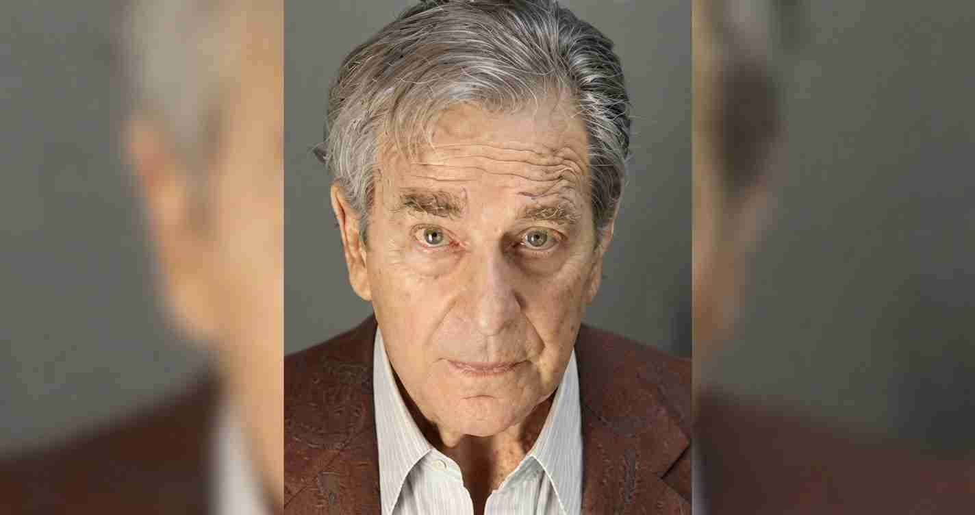 Close-up portrait of an elderly man with gray hair, wearing a brown blazer and light shirt, looking directly at the camera with a serious expression.
