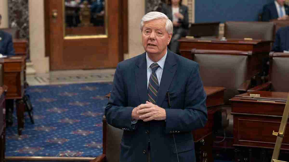 Man in a dark suit speaking in a formal legislative chamber with wooden desks and blue carpetarea behind him, hands clasped together.