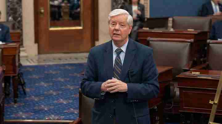 Man in a dark suit speaking in a formal legislative chamber with wooden desks and blue carpetarea behind him, hands clasped together.