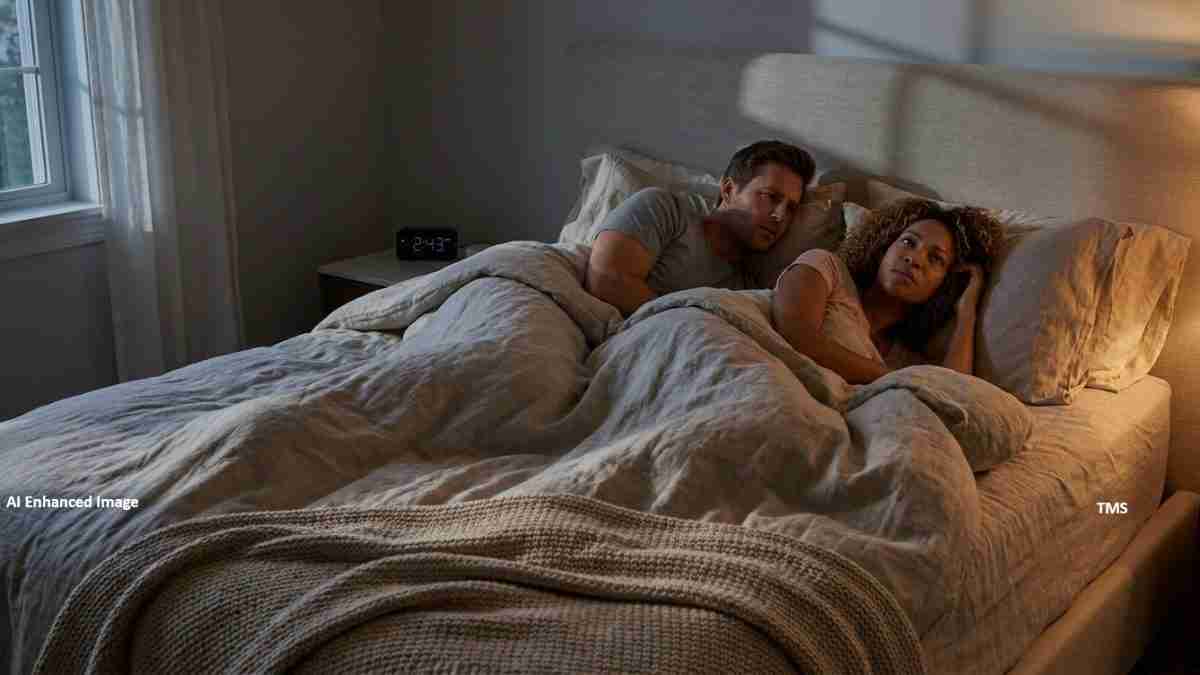Man and woman lying in bed under a gray blanket, awake in a dim bedroom with a window to the left and a clock showing 2:43 on the nightstand.