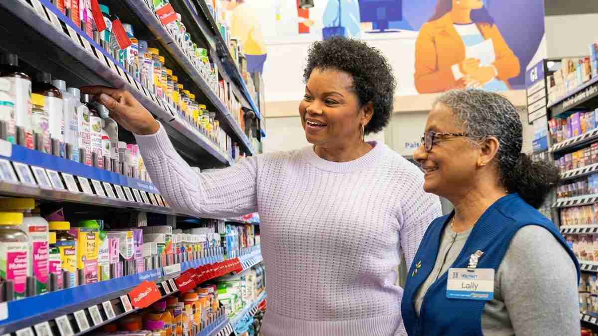 Two women in a store pharmacy aisle; shopper points at products while a staff member in a blue vest smiles and assists.