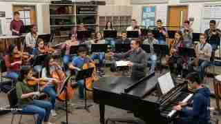 Conductor leads a student orchestra in a rehearsal room with strings, winds, brass, piano, and percussion.